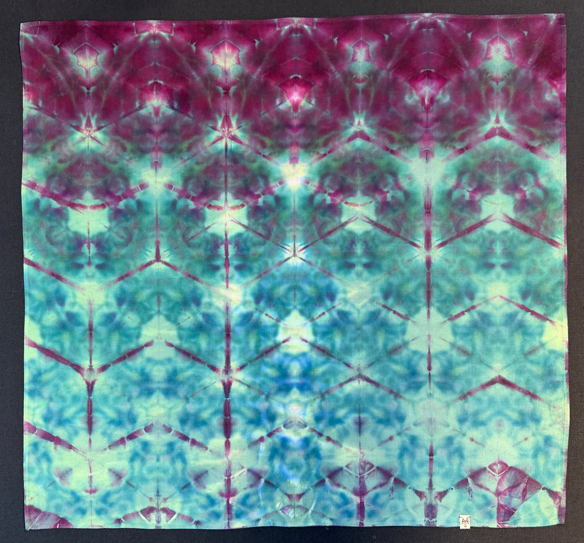 A 100% Cotton Bandana with a symmetrical tie-dye pattern in magenta, teal, aqua, and pale green, photographed flat against a dark background.