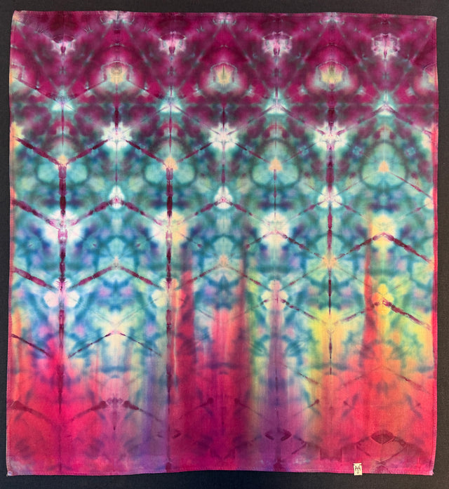 A square tie-dye fabric or textile panel with a symmetrical, kaleidoscopic pattern in magenta, teal, blue, yellow, and red, photographed flat against a dark background.